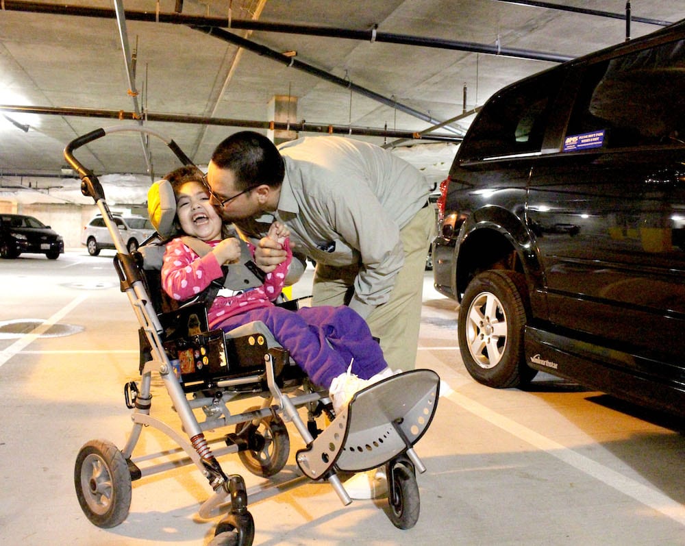 Photo of father kissing his young daughter who uses a wheelchair, they are next to a modified van.