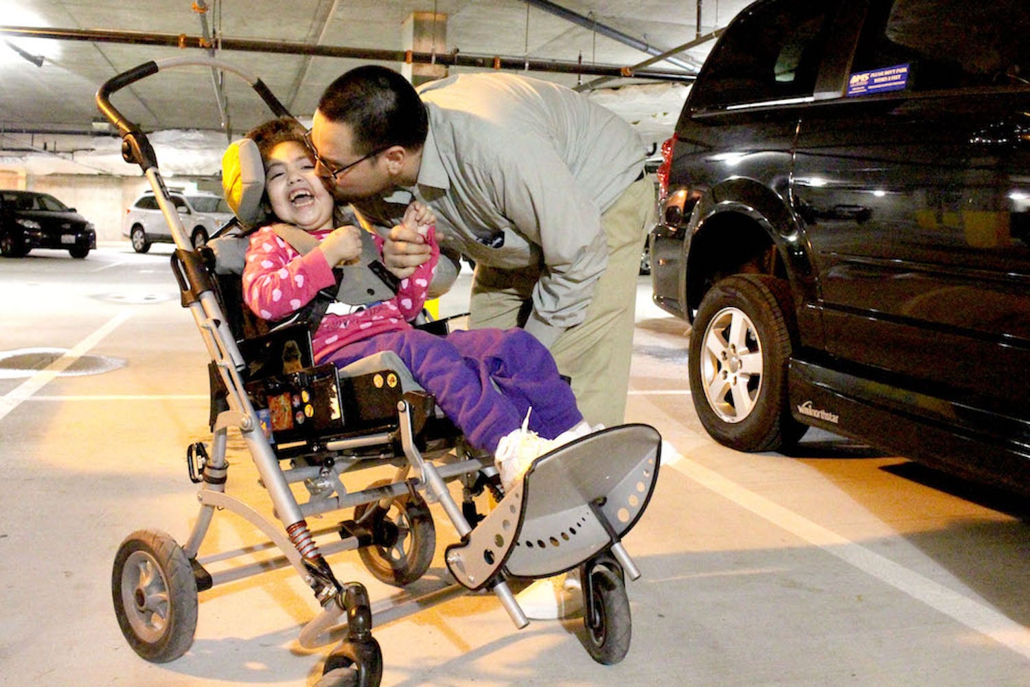 Photo of father kissing the check of his four year old daughter who uses a wheelchair, next to their accessible van, which they were able to purchase through a NWAF Loan