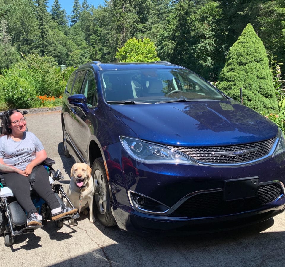 Chelsea sitting in her wheelchair next to her dark blue accessible van. Her dog is sitting next to her.