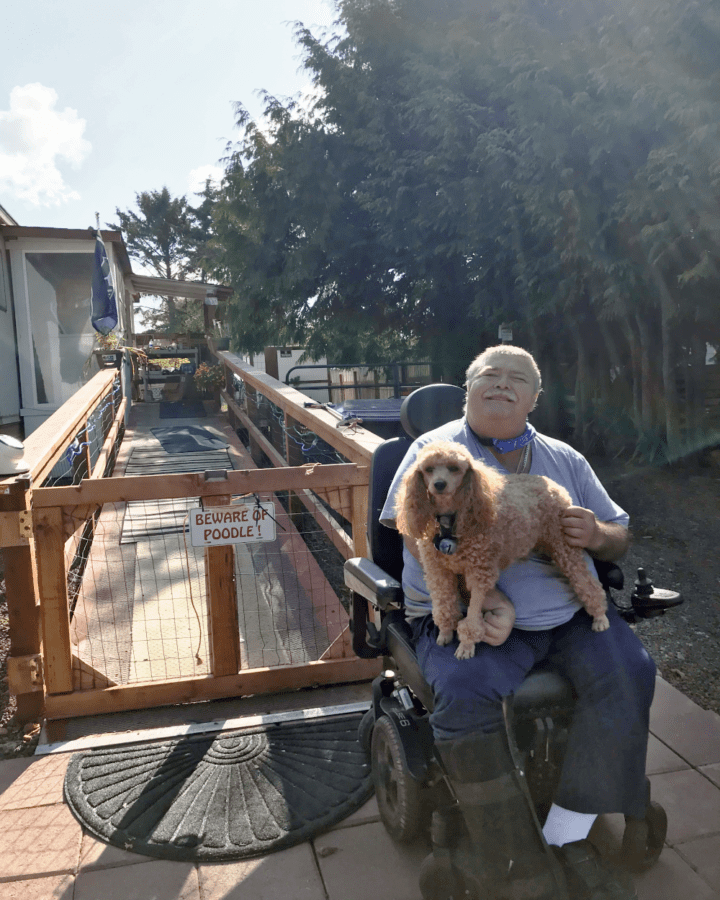 Dave in his wheelchair, sitting in front of the ramp into his house with his poodle on his lap. Behind him, there is a sign that says "Beware of poodle".