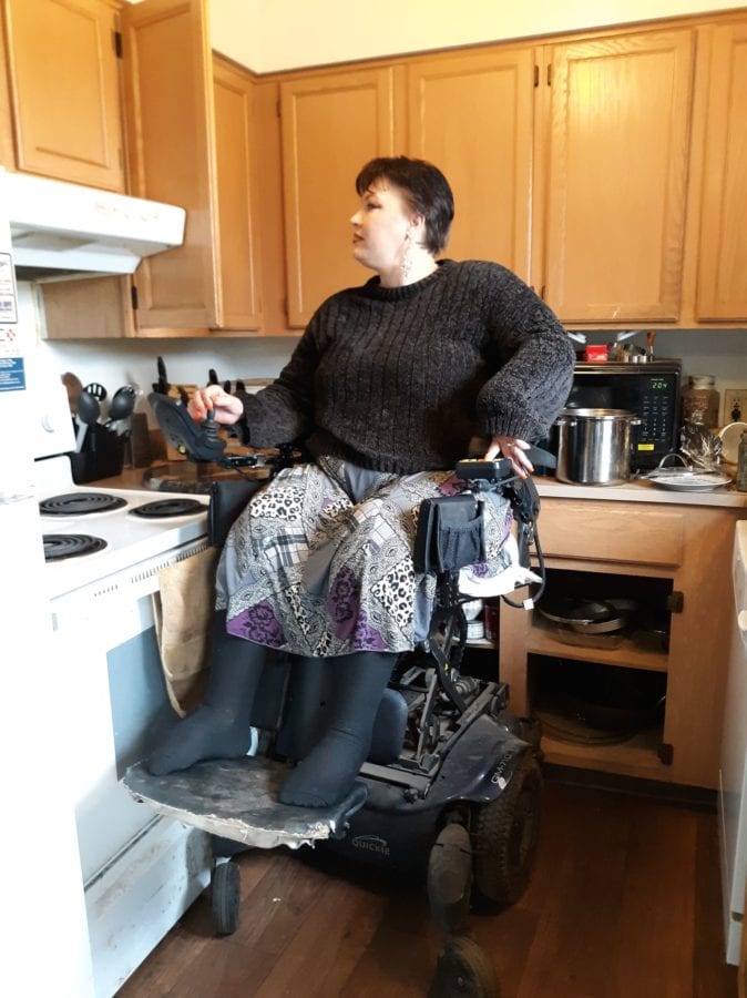 Harley wearing a black sweater and grey skirt, sitting in her wheelchair with the elevating seat lifting her up so that she can look into her cabinet. The cabinet door is open and she is looking in.