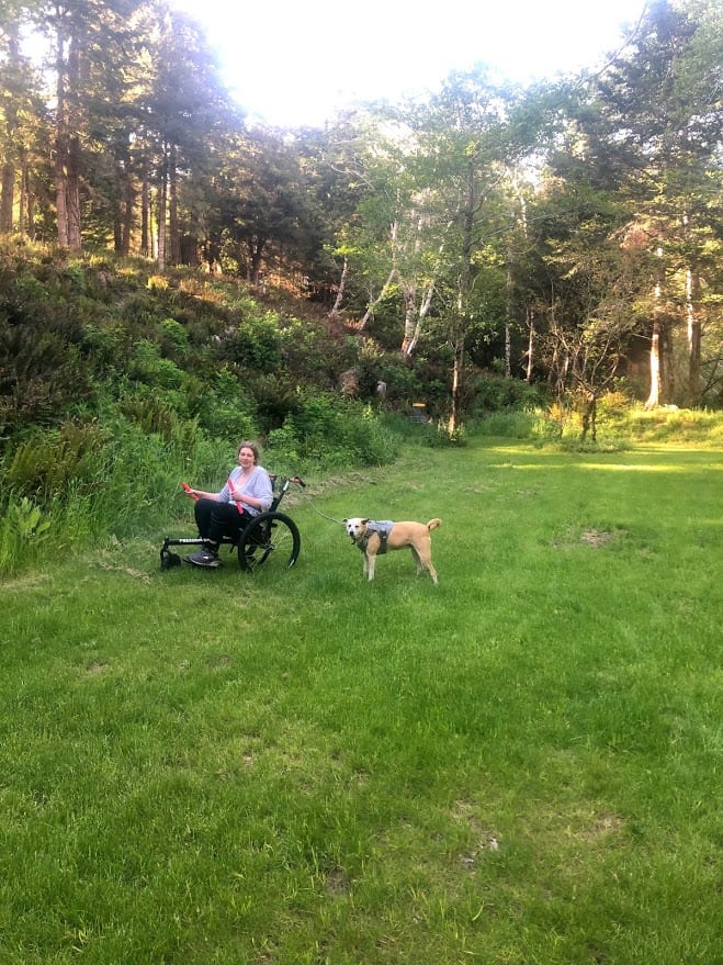 An image of Beth in her chair, walking her dog. She is on grass.