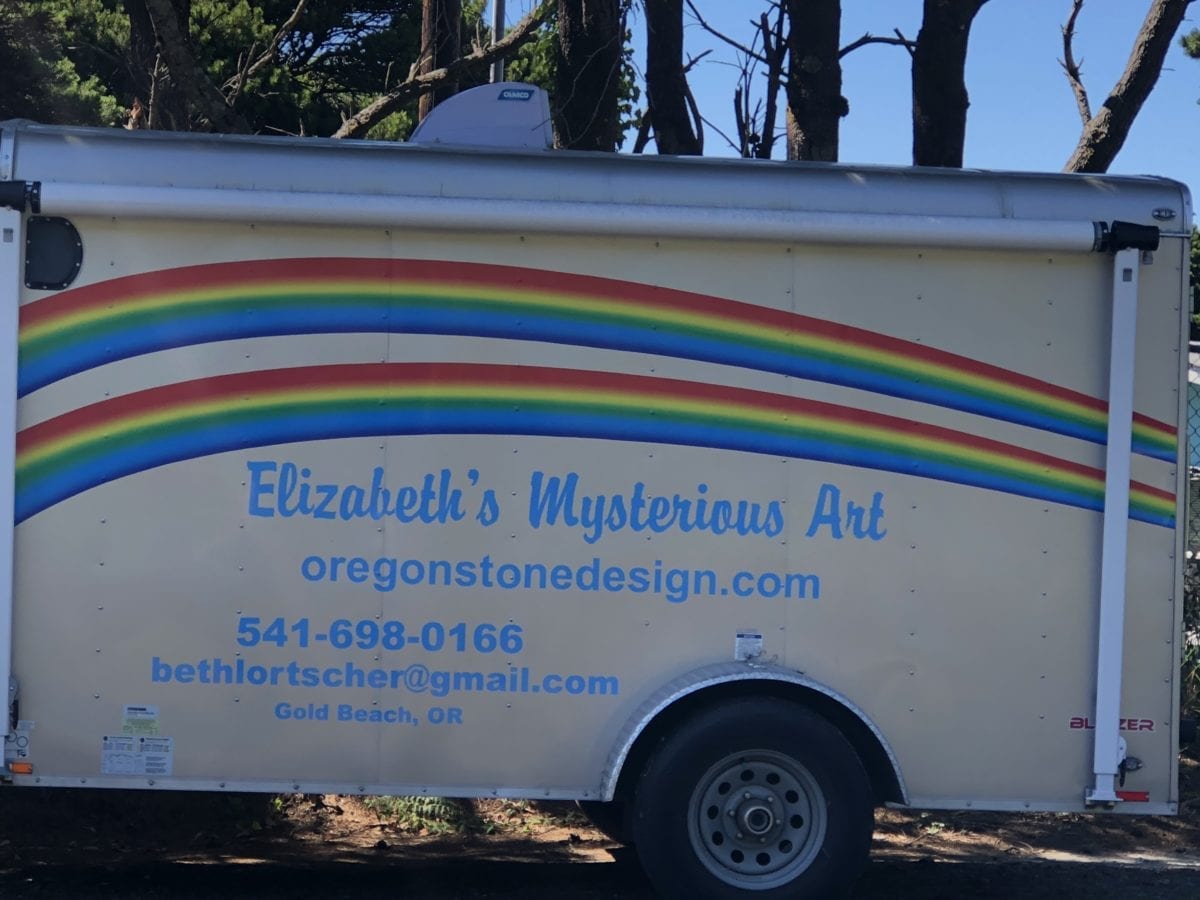 An image of Beth's art trailer, a white trailer with rainbow painted on the side. It says "Elizabeth's Mysterious Art" as well.