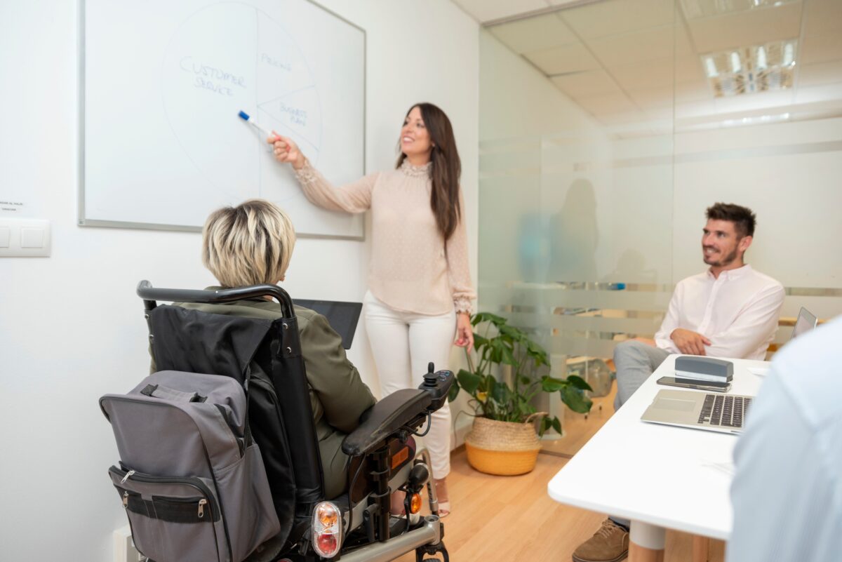 Setting of business meeting. Picture is of a woman using wheelchair looking at another woman pointing to something on a white board.